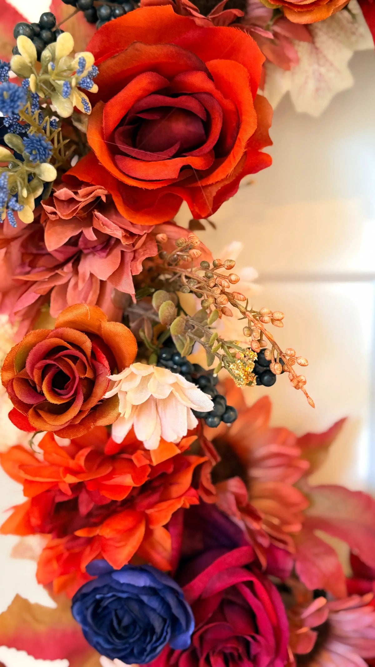 Vibrant fall wreath featuring orange roses, deep blue blooms, berries, and autumn leaves in rich warm tones.
