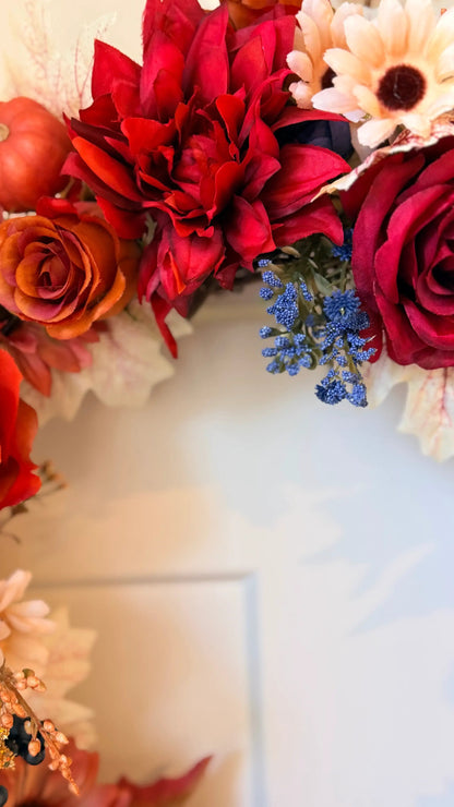 Vibrant fall wreath featuring red dahlias, orange roses, blue accents, and autumn leaves on a white door.
