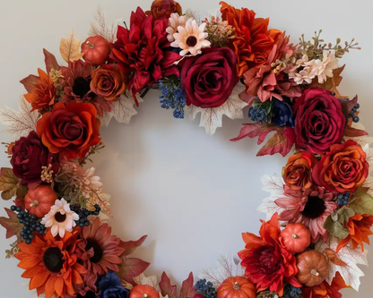 Autumn wreath featuring red roses, orange sunflowers, small pumpkins, maple leaves, and blue berries for fall decor.