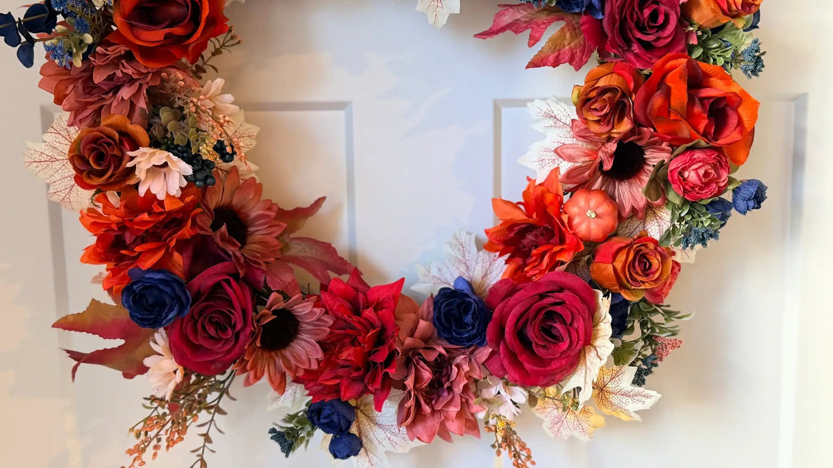 Vibrant fall wreath with red, orange, and blue artificial roses, sunflowers, maple leaves, and a small pumpkin on a white door.