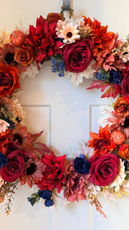 Vibrant fall wreath featuring deep red roses, orange dahlias, blue accents, and small pumpkins on a white door.