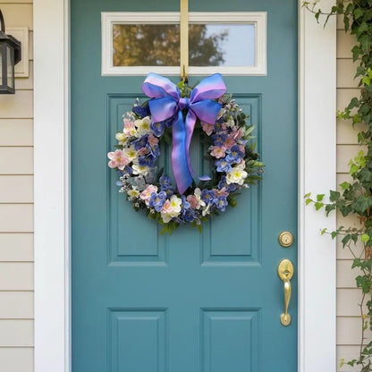 May include: A circular floral wreath featuring a mix of blue, pink, and white blossoms with green foliage, accented by a large, ombre-style bow in shades of purple and blue, hanging on a teal-colored door with a gold handle and a glass window pane.